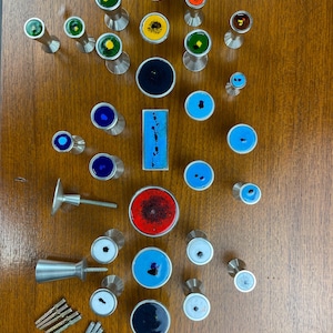 Peut inclure: Assortiment de boutons de cabinet colorés avec différents motifs et designs. Les boutons sont en métal et ont une finition polie. Les boutons sont disposés en grille sur une surface en bois.