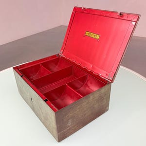 Puede incluir: Una caja de caudales metálica rectangular con un exterior marrón desgastado y un interior rojo vibrante. La tapa está abierta, revelando tres compartimentos internos. Una pequeña placa rectangular en el interior de la tapa muestra el texto Beaumont.