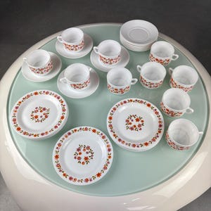 Puede incluir: Un juego de té vintage de porcelana blanca con motivos florales en rojo, naranja y rosa. El juego incluye tazas, platillos y platos, dispuestos sobre una superficie verde claro. Las tazas y los platos tienen un delicado diseño floral.
