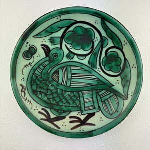 Op de afbeelding: Een ronde, keramische decoratieve schaal met een handgeschilderd vogelontwerp. De schaal heeft een groen en zwart kleurenschema, waarbij het lichaam van de vogel is gedetailleerd met schubben en strepen. De rand en de omtrek van de vogel zijn zwart en de achtergrond is lichtgroen.
