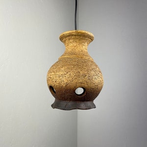 Op de afbeelding: Een bruine en beige keramische hanglamp met een getextureerd oppervlak en gaten rond de onderkant. De hanglamp hangt aan een zwart snoer.