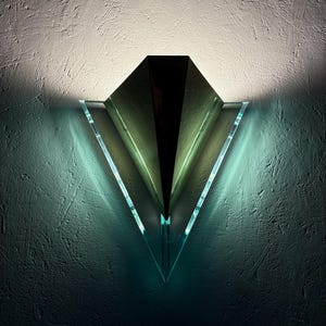May include: Modern wall sconce featuring a black central element and clear, triangular glass panels. The sconce casts a soft, green light, producing a striking effect against a textured wall. The design is contemporary and artistic.