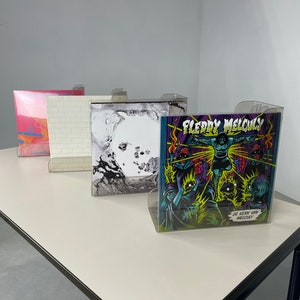 Pu&ograve; includere: Quattro dischi in vinile esposti in supporti di plastica trasparente su un tavolo bianco. I dischi sono in stili diversi, tra cui un design astratto rosa e arancione, un design a muro di mattoni bianchi, un design marmoreo bianco e nero e un design di cartoni animati colorato con il testo "Fleddy Melculy De Kerk Van Melculy".