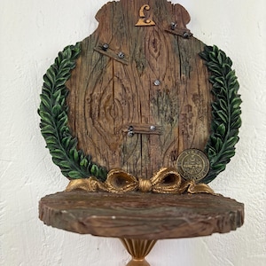 May include: A decorative wooden shelf with a fairy door design. The shelf features a faux wood grain texture, a green laurel wreath, and a gold-colored bow. A small gold letter "L" is visible.
