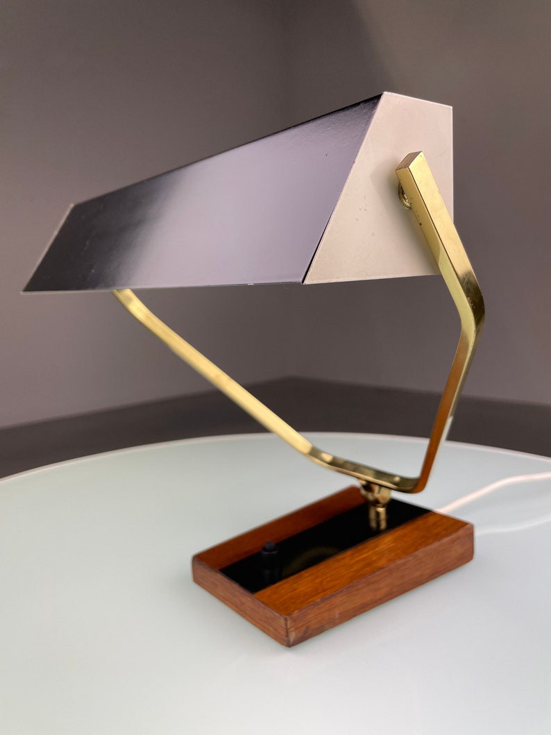 May include: A mid-century modern desk lamp with a brass frame and a black and white metal shade. The lamp has a wooden base and a black switch.