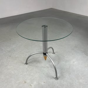 Op de afbeelding: Een ronde glazen tafel met een zilveren metalen onderstel. Het onderstel is gevormd als een potlood met een puntige punt en drie poten.