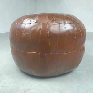 Op de afbeelding: Een ronde bruine leren poef met een gestikt ontwerp. De poef is glad.