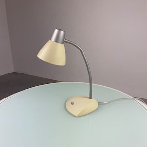 Puede incluir: Una lámpara de escritorio blanca y plateada con un brazo flexible. La lámpara tiene una pantalla blanca y una base plateada.