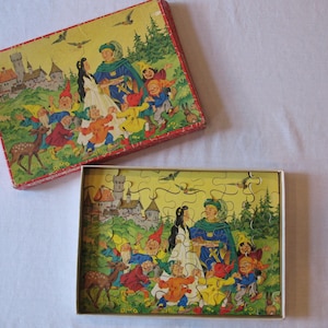 Op de afbeelding: Een vintage puzzel die een sprookjesscène afbeeldt met een prins en een prinses omringd door een groep dwergen. De puzzel zit in een doos met een kleurrijke illustratie van dezelfde scène.