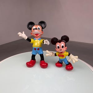 May include: Two plastic Mickey Mouse figurines, one larger than the other. The larger Mickey Mouse is wearing a yellow shirt, blue shorts, and red shoes. The smaller Mickey Mouse is wearing a yellow shirt, blue shorts, a red bow tie, and red shoes.