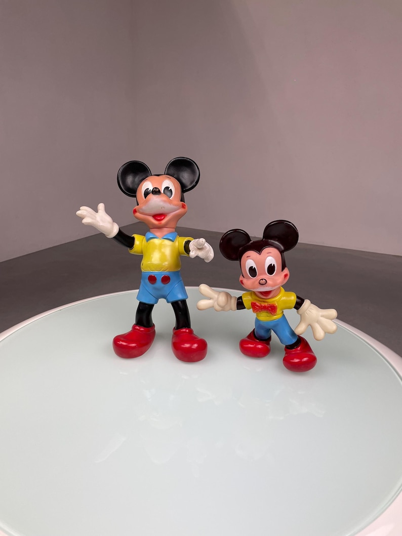 May include: Two plastic Mickey Mouse figurines, one larger than the other. The larger Mickey Mouse is wearing a yellow shirt, blue shorts, and red shoes. The smaller Mickey Mouse is wearing a yellow shirt, blue shorts, a red bow tie, and red shoes.