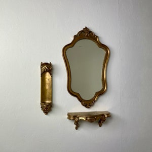 May include: A gold-toned ornate mirror with a shelf underneath. The mirror has a scalloped edge and the shelf has a curved design.