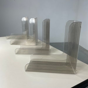 Pu&ograve; includere: Set di quattro porta riviste in plastica trasparente. I porta riviste sono impilati su una superficie bianca. Ogni porta riviste ha un fronte curvo e un retro piatto.