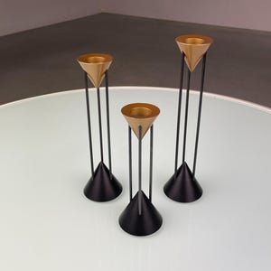 May include: Three gold candle holders with black metal stands. Each holder has a triangular base and three vertical supports.