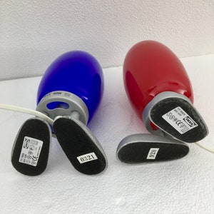 May include: Two table lamps with a blue and red glass shade. The lamps are made of metal and have a silver finish. The base of each lamp has a black label with the text "IKEA" and a model number.