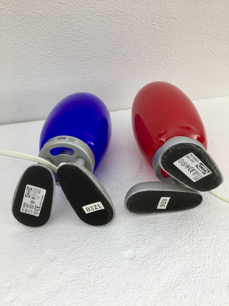 May include: Two table lamps with a blue and red glass shade. The lamps are made of metal and have a silver finish. The base of each lamp has a black label with the text "IKEA" and a model number.