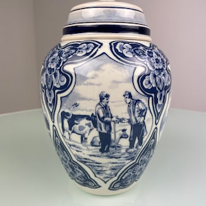 Op de afbeelding: Een witte keramische pot met een blauw-wit bloemenmotief. De pot heeft een deksel en toont een scène met twee mensen, koeien en een varken.