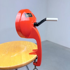 Könnte beinhalten: Ein leuchtend orangefarbenes, manuelles Pede-Küchengerät mit schwarzer Kurbel und weißem Griff. Das Gerät verfügt über einen Klemmmechanismus an der Basis zur Befestigung an einer Oberfläche und ein schwarzes Etikett mit der Aufschrift Pede Made in Holland.