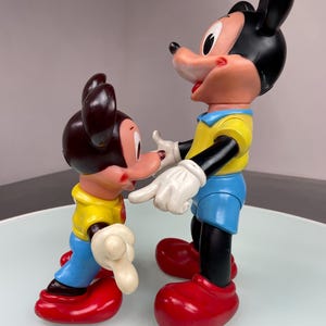 May include: Two vintage plastic Mickey Mouse figures. The larger figure is standing with its arms outstretched, while the smaller figure is standing with its arms at its side. Both figures are wearing yellow shirts, blue pants, and red shoes.