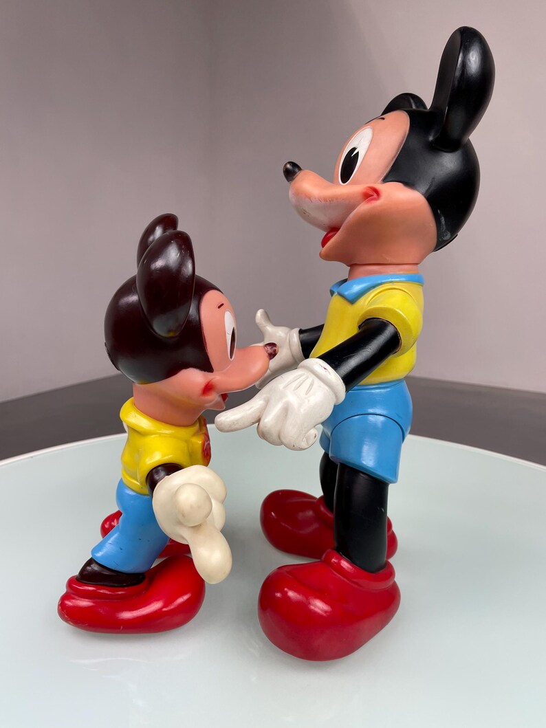May include: Two vintage plastic Mickey Mouse figures. The larger figure is standing with its arms outstretched, while the smaller figure is standing with its arms at its side. Both figures are wearing yellow shirts, blue pants, and red shoes.