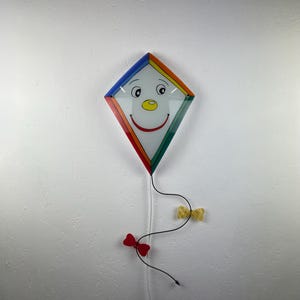 Può includere: Un aquilone di vetro colorato con un viso sorridente, appeso a un cordino con fiocchi rossi e gialli.