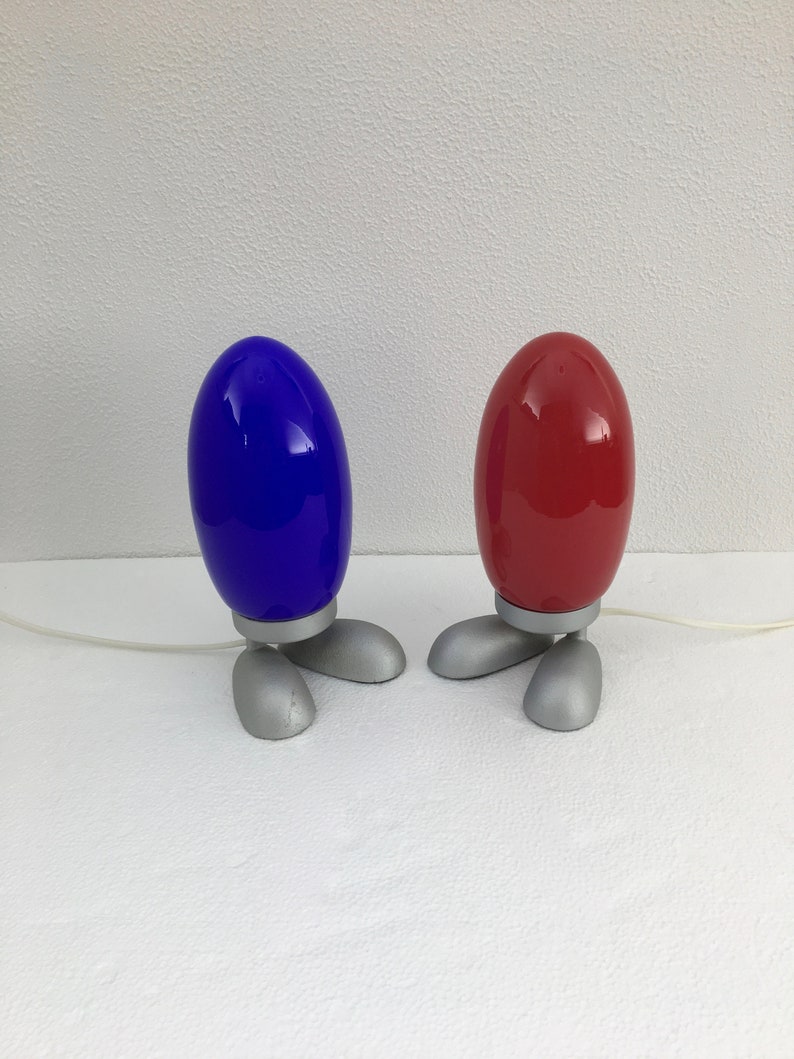 May include: Two table lamps with a unique design. The lamps are shaped like eggs with three legs. One lamp is blue and the other is red.
