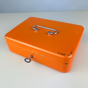 Op de afbeelding: Een rechthoekige oranje metalen geldkist met een zilverkleurig metalen handvat op het deksel. De kist heeft een sleutel in het slot aan de voorzijde. De oranje lak vertoont lichte gebruikssporen en slijtage langs de randen.