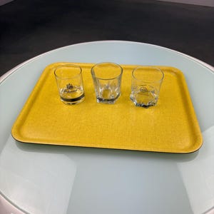 Op de afbeelding: Drie heldere borrelglaasjes op een gele rechthoekige dienblad. De dienblad staat op een wit en lichtblauwe tafel. Eén borrelglaasje heeft een zwart en goud logo. De andere twee borrelglaasjes zijn effen.