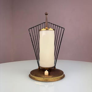 Könnte beinhalten: Eine Tischleuchte mit einem zylindrischen weißen Glasschirm, der in einem schwarzen, fächerförmigen Drahtgestell zentriert ist. Die Leuchte steht auf einem runden Messingsockel mit einem kleinen weißen Druckschalter in der Mitte.