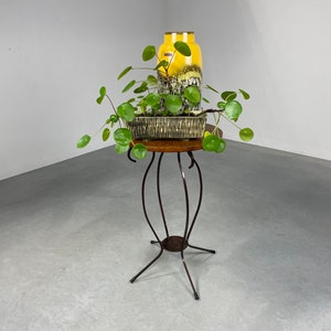 Op de afbeelding: Een gele en bruine keramische vaas met een groene plant in een zwart-witte pot op een ronde houten tafel met een zwarte metalen basis.