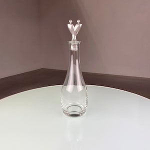 May include: A clear glass decanter with a silver stopper featuring two stylized figures. The stopper is shaped like a heart.