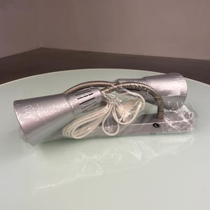 Può includere: Una lampada da scrivania argentata con due bracci flessibili a collo di cigno e un cavo bianco. La lampada è confezionata in plastica.