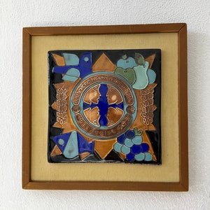 Op de afbeelding: Een ingelijste keramische tegel met een blauw en gouden ontwerp. De tegel heeft een kruis, een vogel, druiven, tarwe en een appel. De tekst "Katholieke vrouwen organisatie 1933 Zeelst 1963" staat rond het kruis.