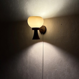 Op de afbeelding: Een wandlamp met een ronde, gebroken witte glazen kap. De armatuur heeft een donkerbruine basis en een trekschakelaar. Het licht werpt een warme gloed op de getextureerde muur.