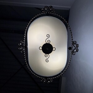 Op de afbeelding: Een ovale plafondlamp met een crèmekleurige geplooide kap omringd door een donker, gedraaid metalen frame. Het midden van de lamp heeft een zwart cirkelvormig accent met decoratieve kruldetails die zich aan de boven- en onderkant uitstrekken.