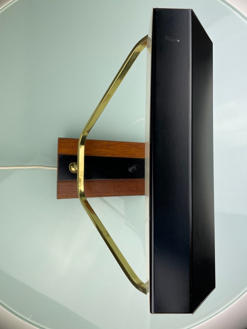 May include: A black and brass wall mounted lamp with a rectangular shade. The lamp has a wooden base with a brass arm.