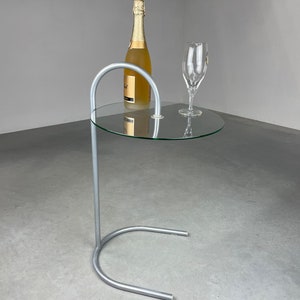 Op de afbeelding: Een zilveren metalen bijzettafel met een rond glazen blad. Op de tafel staan een fles champagne en een wijnglas.