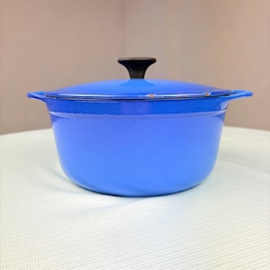 May include: A bright blue enameled cast iron Dutch oven with a matching lid and a black knob handle. The pot features two integrated side handles and shows minor signs of wear along the rim of the lid, revealing a metallic base layer.