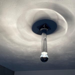 Może przedstawiać: Nowoczesna lampa sufitowa z czarną kwadratową podstawą i długim, cylindrycznym, srebrnym kloszem. Żarówka jest przezroczysta, okrągła i ma srebrne wykończenie.