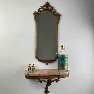 Könnte beinhalten: Ein antiker goldgerahmter Spiegel hängt über einem kleinen Regal mit Marmorplatte. Auf dem Regal stehen kleine Glasflaschen mit roten Verschlüssen und eine große Flasche mit türkisfarbenem Etikett. Spiegel und Regal sind an einer weißen Wand befestigt.