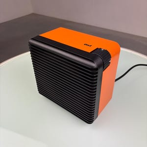 Peut inclure: Radiateur électrique orange et noir avec un panneau avant à nervures et un cadran sur le côté. La marque "ewt" est imprimée sur le dessus. Un cordon d'alimentation noir s'étend à l'arrière.