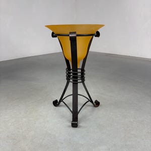 Peut inclure: Un support décoratif composé d'un bol conique en verre dépoli ambré maintenu par une structure en métal sombre. Le cadre comprend trois supports verticaux, un accent central en métal enroulé et une base tripode aux pieds incurvés.