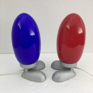 May include: Two table lamps with a unique design. The lamps have a round, egg-shaped shade in blue and red. The base of each lamp is silver and has three feet.