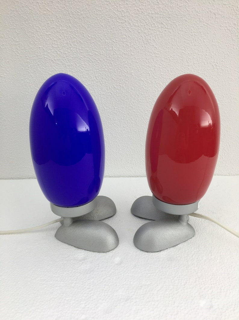 May include: Two table lamps with a unique design. The lamps have a round, egg-shaped shade in blue and red. The base of each lamp is silver and has three feet.