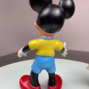 May include: A vintage plastic Mickey Mouse figurine wearing a yellow shirt, blue shorts, and red shoes. The figurine is standing on a white surface.