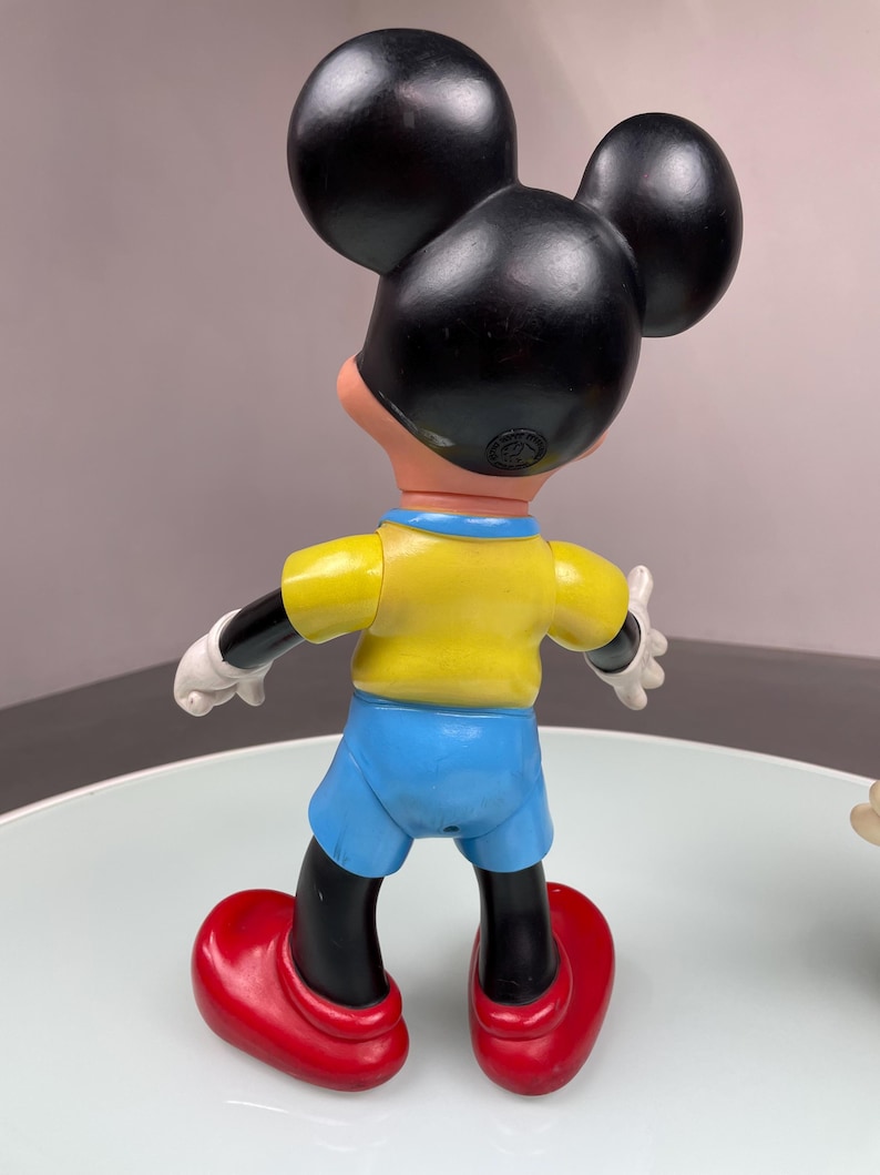 May include: A vintage plastic Mickey Mouse figurine wearing a yellow shirt, blue shorts, and red shoes. The figurine is standing on a white surface.