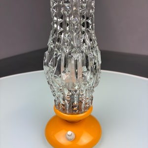 Op de afbeelding: Een vintage tafellamp met een heldere, getextureerde glazen kap en een oranje voet. De kap heeft een geometrisch patroon en de voet heeft een rond ontwerp met een kleine schakelaar. Decoratieve verlichtingsarmatuur.