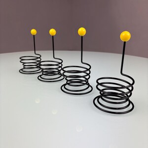 May include: Four black metal spiral holders with yellow ball tops. The holders are arranged in a row, with the yellow balls at the top. The holders are on a white surface.
