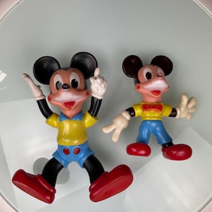 May include: Two vintage plastic Mickey Mouse figurines. The larger figurine is standing with its arms raised and wearing a yellow shirt, blue shorts, and red shoes. The smaller figurine is standing with its arms out to the sides and wearing a yellow shirt, blue pants, and red shoes.