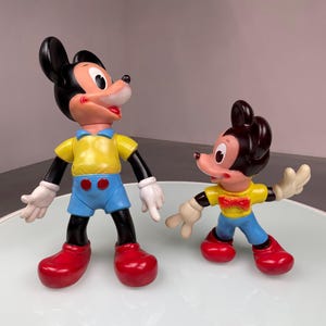 May include: Two plastic Mickey Mouse figurines. The larger figurine is wearing a yellow shirt, blue shorts, and red shoes. The smaller figurine is wearing a yellow shirt with a bow tie, blue pants, and red shoes.
